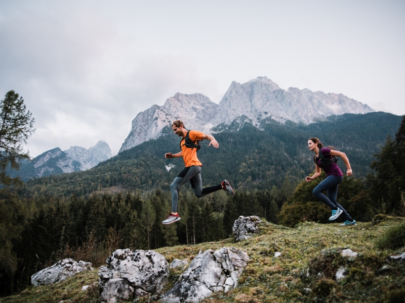 Zwei Trailrunner stürmen über einen felsigen Weg vor Bergpanorama